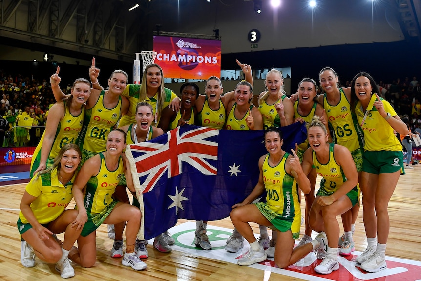 A netball team gathers on the court for a group photo holding an Australian flag as a sign reads 