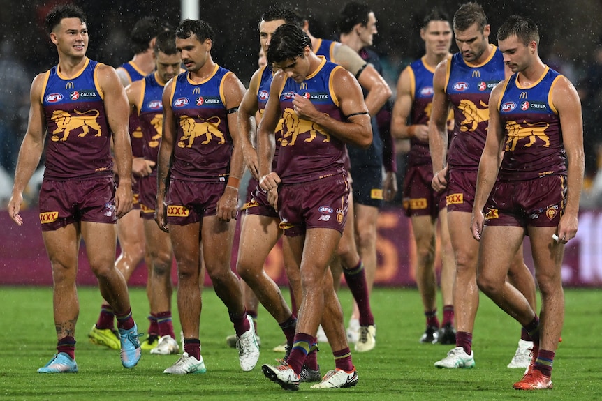 A group of dejected Brisbane Lions AFL players rise from the ground with their heads bowed.