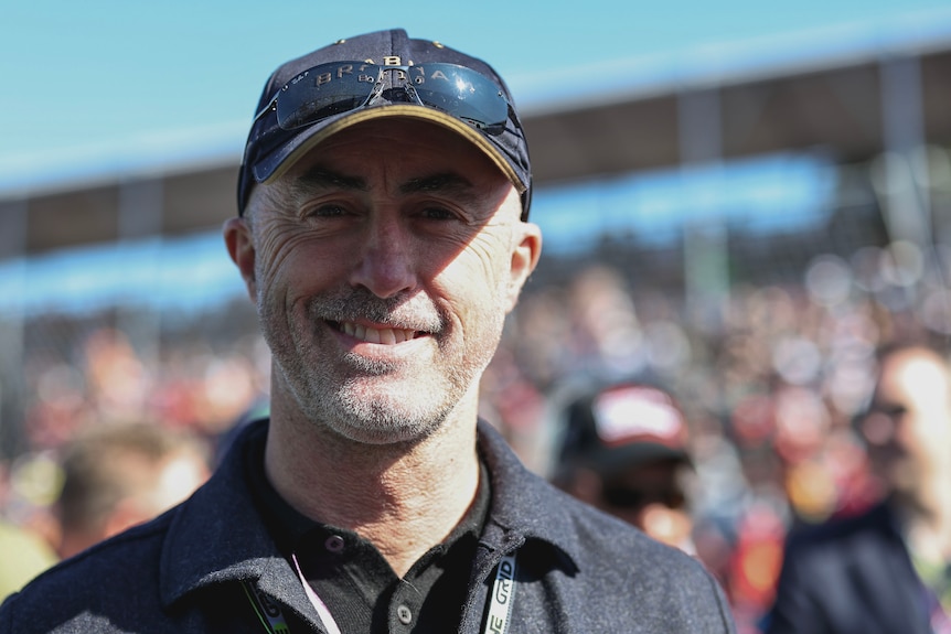Former F1 driver David Brabham, wearing a black cap, smiling for a photo while standing on an F1 grid.