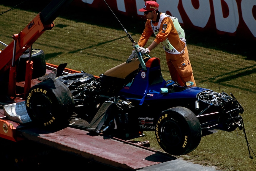 The remains of a wrecked F1 car are lifted onto a crane, with the help of a track marshal