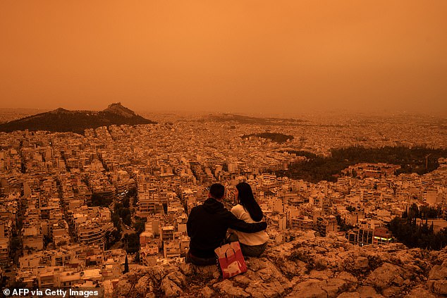 A couple pictured sitting on the hill looking out over a Mars-like Athens after southerly winds painted the city an unusual hue.
