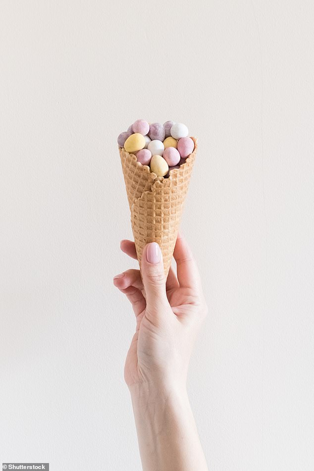 A woman's hand holding a sweet waffle ice cream cone decorated with mini Easter chocolate eggs on a light pastel background (stock image)
