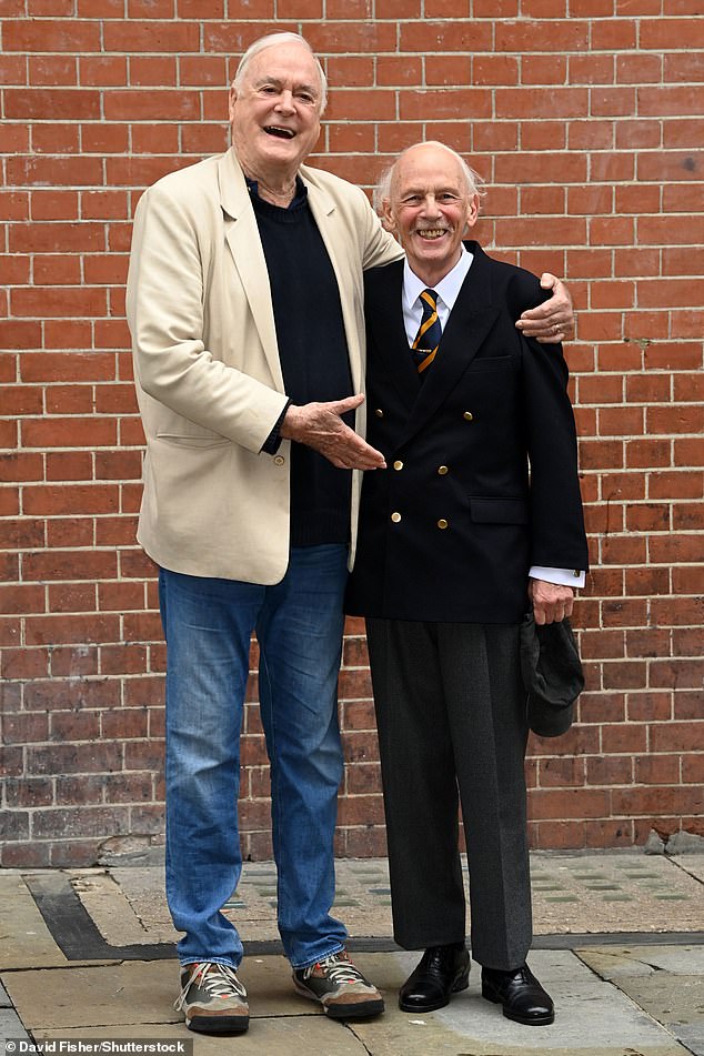 Cleese and Nicholas posing together and smiling despite the bad weather.  Nicholas donned a smart suit with gray pants and a black double-breasted jacket with gold buttons to complement his gold and blue tie.
