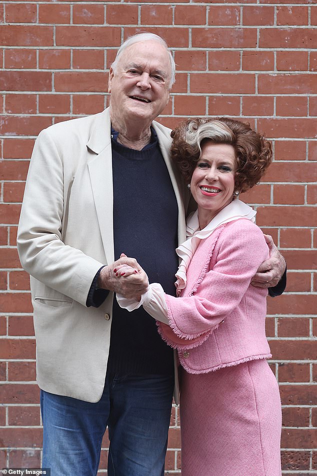 Cleese posing with Anna-Jane Casey, who was wearing her character Polly's iconic outfit: a pink suit and a ruffled white blouse with her curly hair tied up on top of her head.