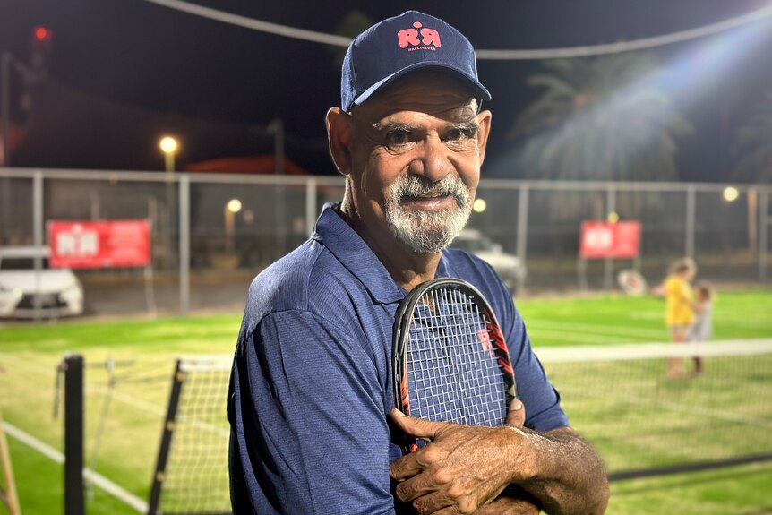 A man wearing a baseball cap holds a tennis racket against his chest under the lights of a tennis court at night. 