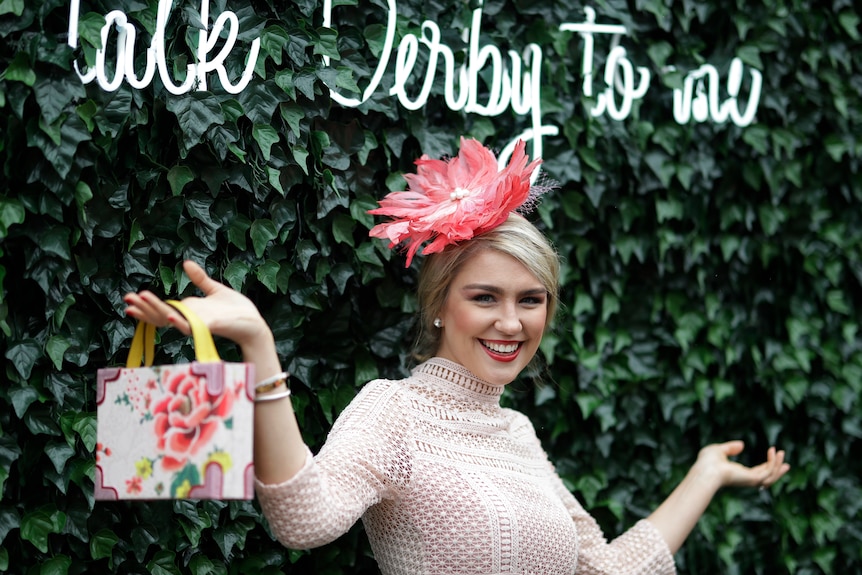 A woman, wearing a headdress in front of a garden wall, at a horse race, holding a purse