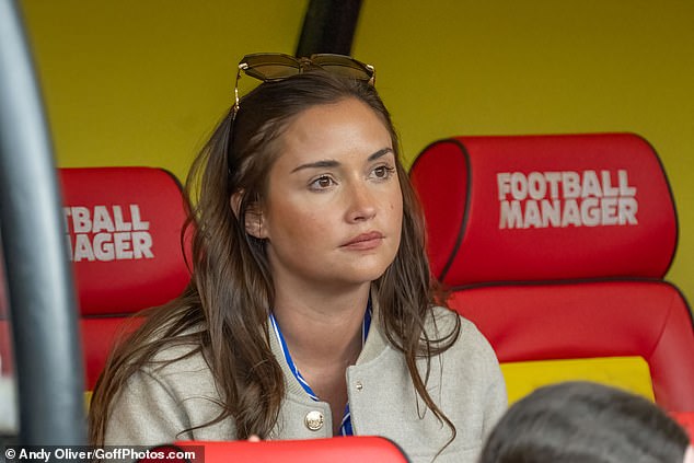 A tanned Jacqueline stunned in a tan baseball jacket worn over a blue and white striped dress.