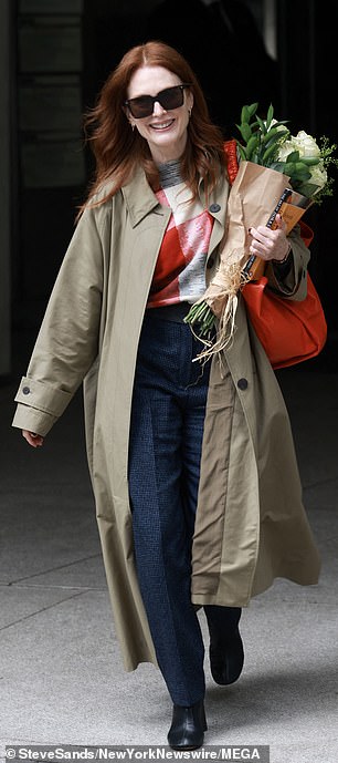 Moore carried an orange purse and wore a classic khaki trench coat over a colorful printed blouse, dark rinse denim, and black high-heeled ankle boots.