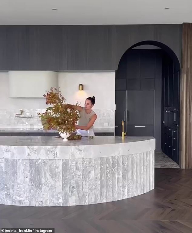 Jesinta shared a video of herself setting up a floral arrangement in the stone kitchen, which features a curved island bench, all-black cabinets, and a pantry.