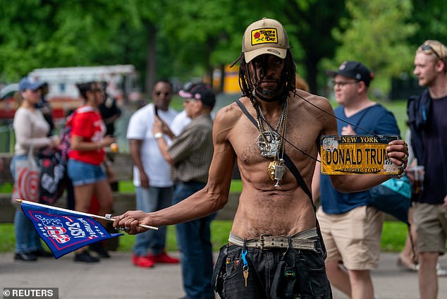 A shirtless Trump supporter wearing jeans, holding a flag and a Trump badge walks outside the rally.