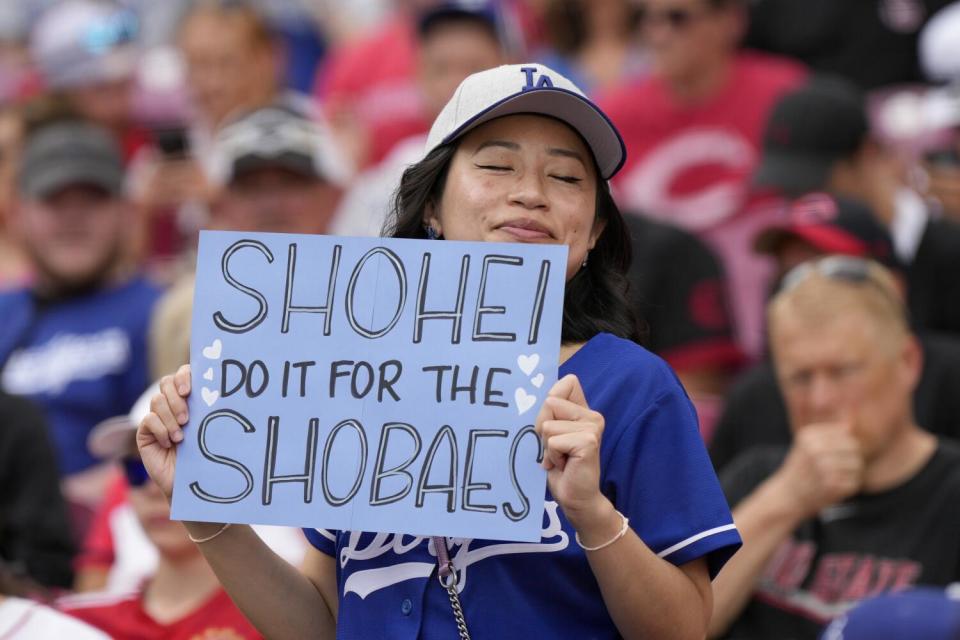 An Ohtani fan wearing a Dodger cap and jersey holds a sign that reads "Shohei do it for the Shoebaes" at the Great American Ballpark.