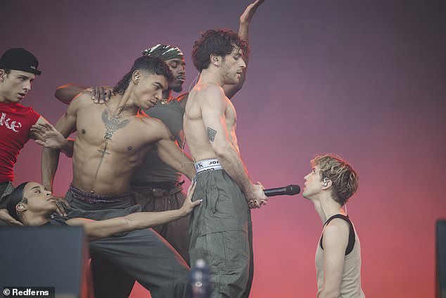 Troye kneels on the floor while one of his dancers holds the microphone in an X-rated position. Troye sings, 'Boy, can I be honest?' before recovering the microphone