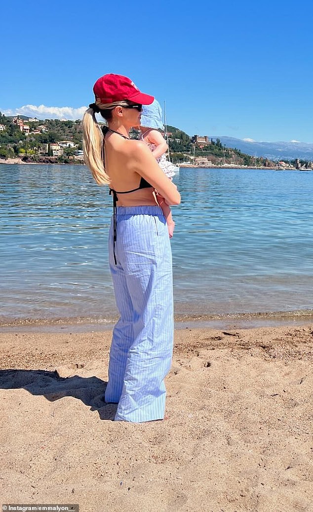 Wearing a black bikini, long blue pinstriped pants and a red baseball cap, the wife of cricket star Nathan Lyon posed with their baby on the shore of a cove