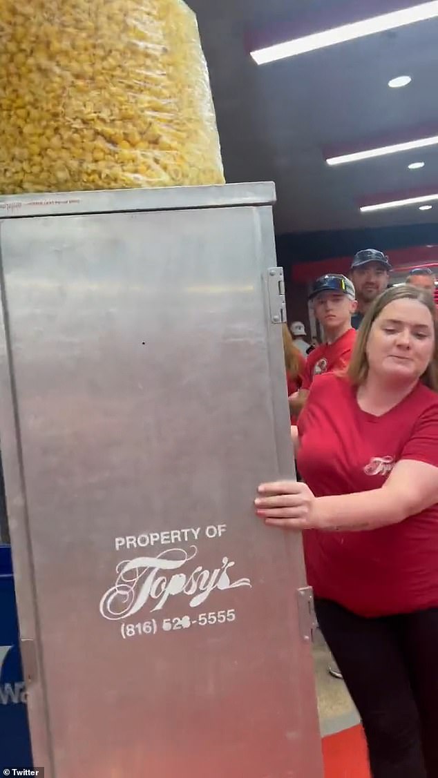 A woman dressed in red with her lips pursed, pushing a large metal container with a bag of popcorn perched precariously on top.