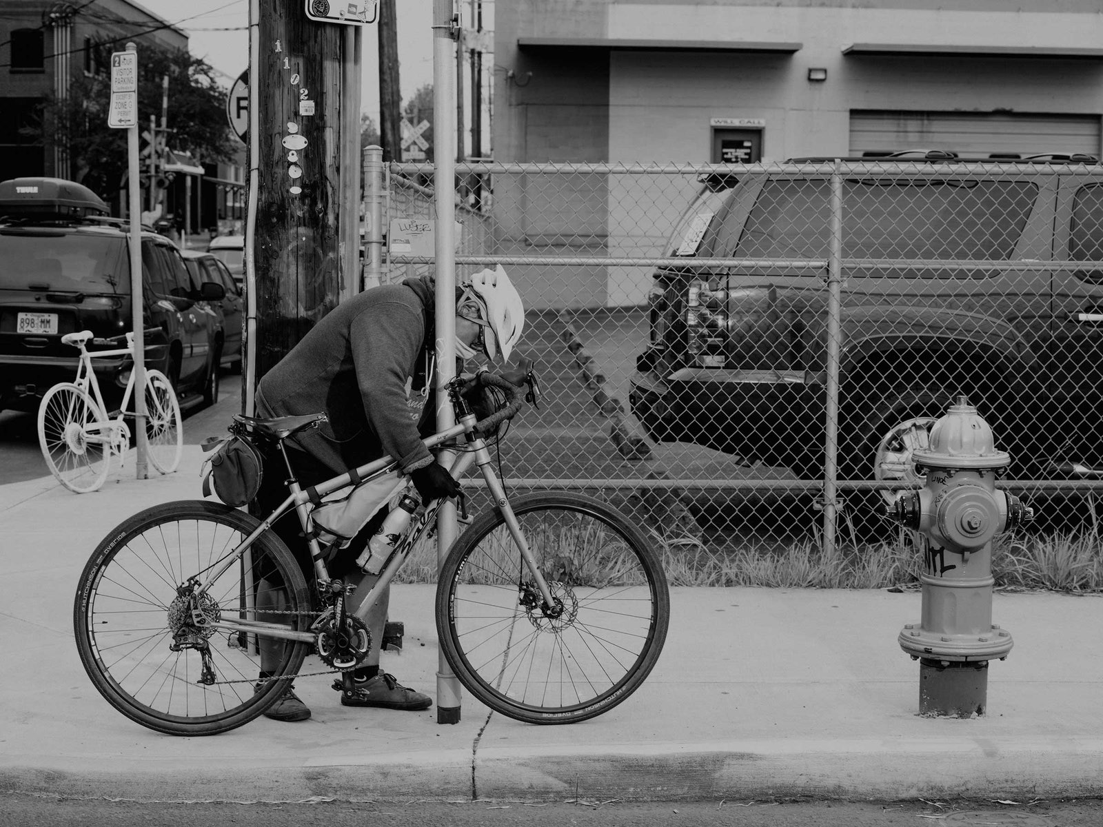 A black and white photograph of a person chaining a bicycle to a street sign with cars and buildings in the background.