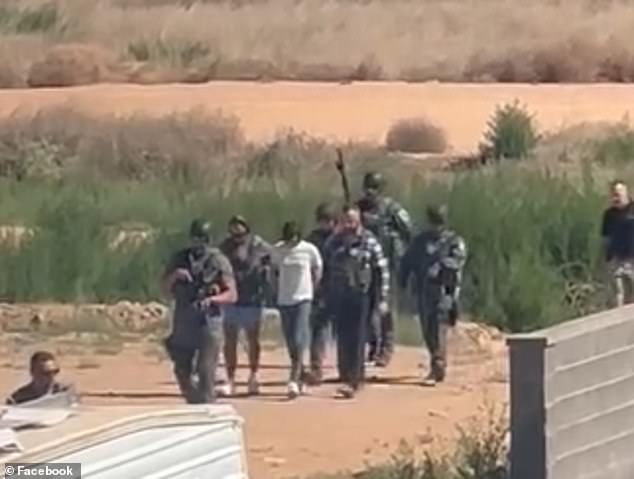 Flanked by seven officers, some of whom are wearing tactical gear and carrying assault rifles and a dog, Bailey is led from a desert area with two men restraining her.