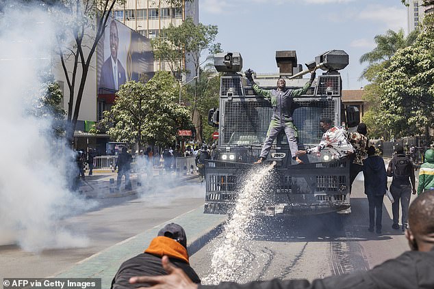 Protesters are perched on a police water cannon attempting to repel them with chemically treated water jets next to an advertisement displaying a photograph of Kenyan President William Ruto.