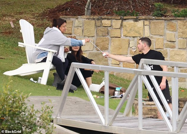 Nick was seen handing a skewer to someone sitting in a white Adirondack chair, presumably preparing to toast marshmallows.