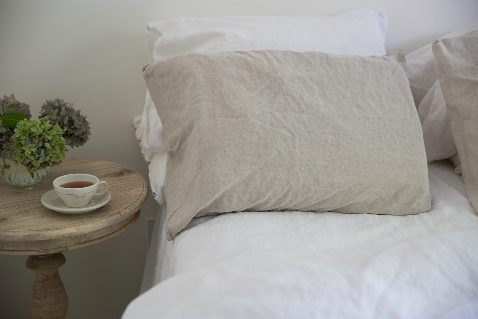 Bed made with white linen and a side table with a vase of hydrangeas and a cup of tea.