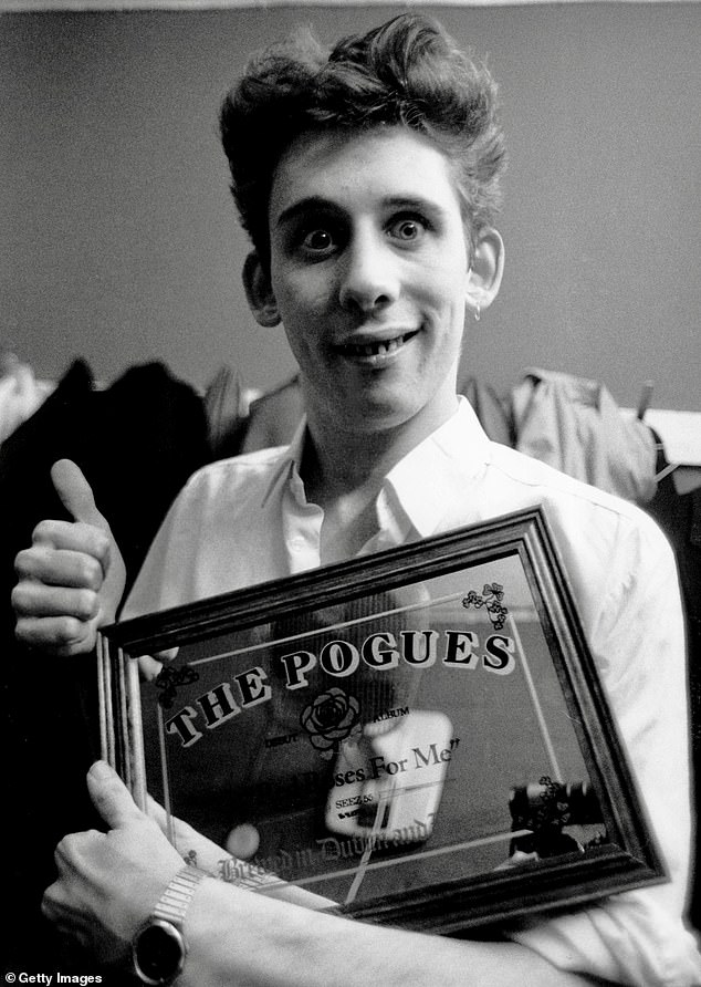 Pictured: Shane MacGowan in 1984 holding a mirrored plaque referencing the band's 1984 album 'Red Roses for Me'.