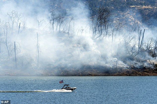 A boat crosses Lake Oroville with a smoldering hillside behind as the Thompson Fire burns in Oroville, California, Wednesday, July 3, 2024.