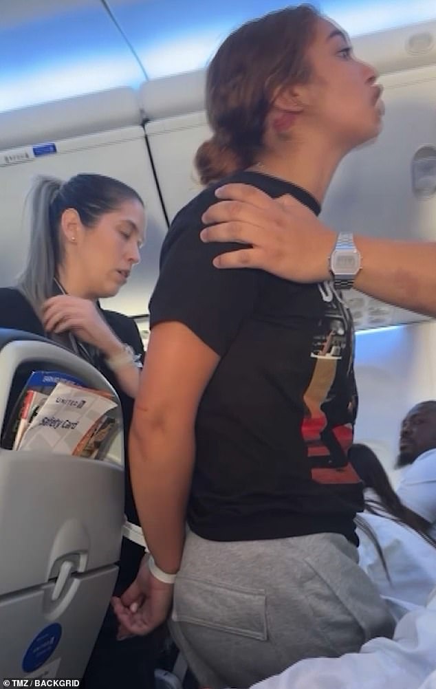 The video shows the woman, now with her hands tied behind her back and standing in the aisle, yelling at everyone not to fly United.