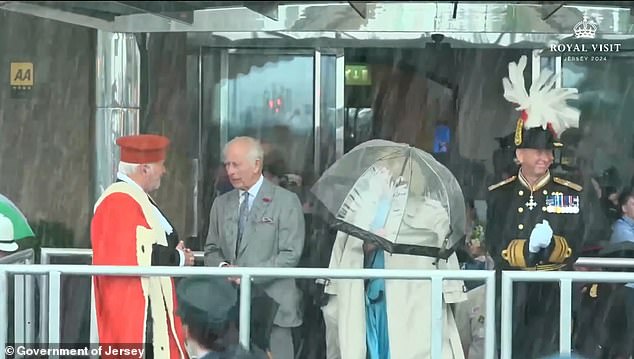 King Charles shouted to his aide to help Queen Camilla as she struggled to put on her coat while battling wind and rain during engagements in Jersey on Monday.