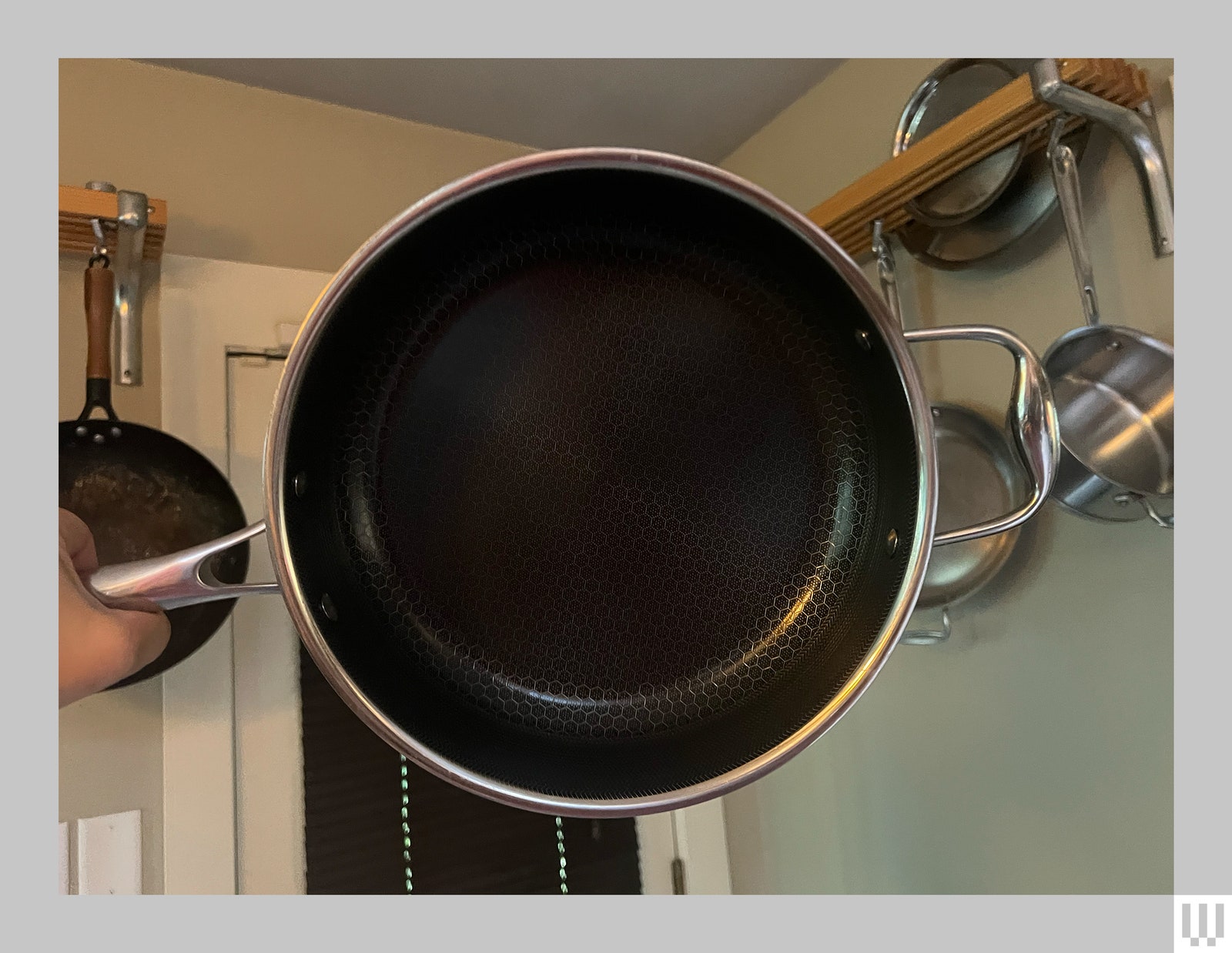 Hand holding a silver frying pan with a door and a shelf with other pans in the background