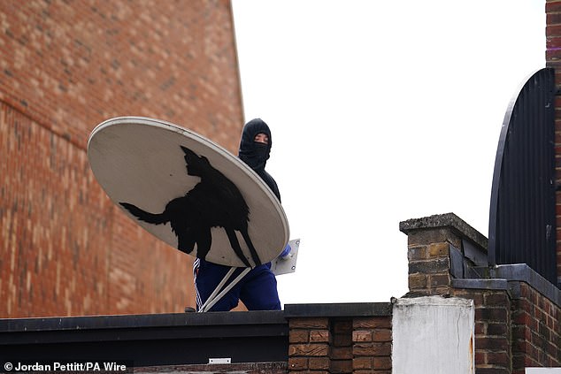 A hooded man wearing a mask holding the satellite dish while standing on top of the building.