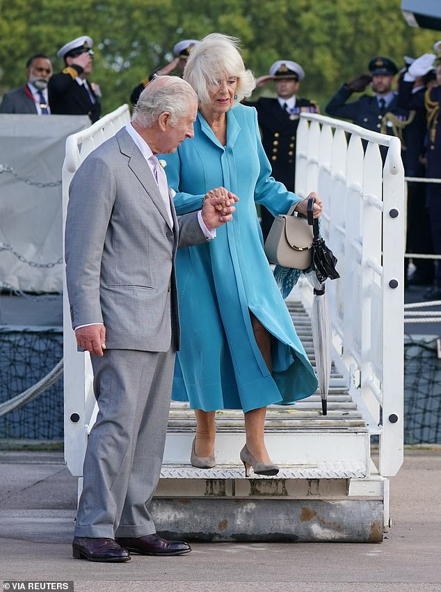 The King helps Camilla, who is wearing a long blue coat and carrying a beige handbag, off the flight deck of HMS Iron Duke in Bordeaux during the couple's state visit to France, September 2023.