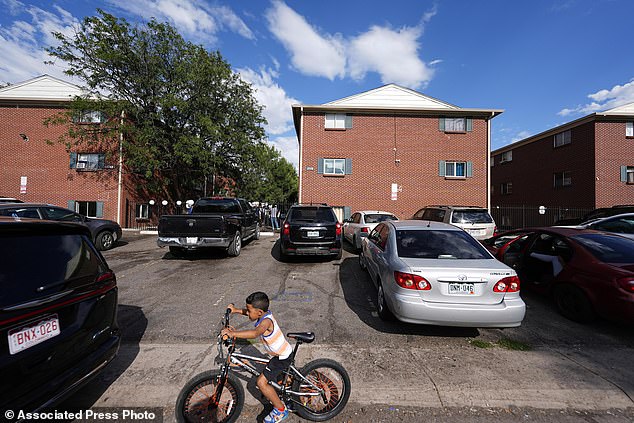 A boy rides his bicycle between apartment buildings as a demonstration takes place in the courtyard to address chronic problems in apartment buildings occupied by people displaced from their home countries in Central and South America in Aurora.