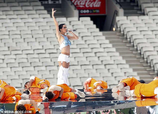 Katy is up front, singing into the microphone, wearing her metallic blue top and white shorts with attached billowing leg covers, continuing the futuristic theme.