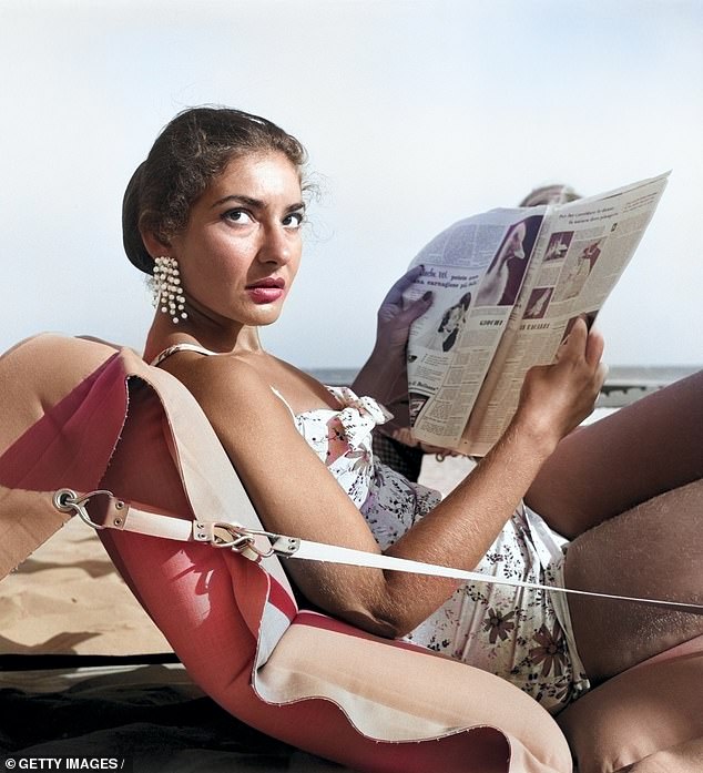 American-born Greek opera singer Maria Callas, lying on an inflatable mattress on the beach at Lido di Venezia, wearing a floral bathing suit and dangling earrings, reading a newspaper, Venice, 1950