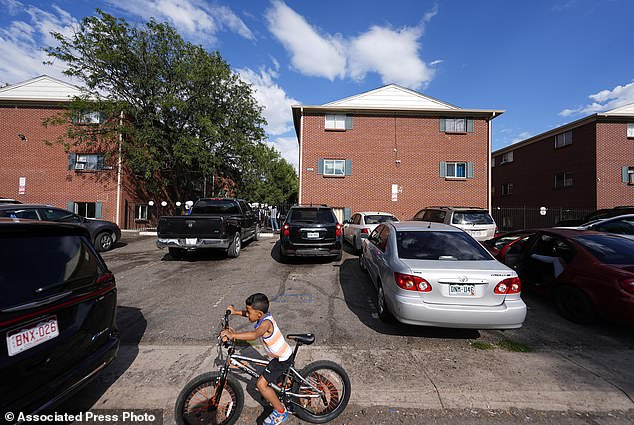 A boy guides his bicycle past apartment buildings as a courtyard demonstration takes place to address chronic problems in apartment buildings occupied by people displaced from their home countries in Central and South America in Aurora.