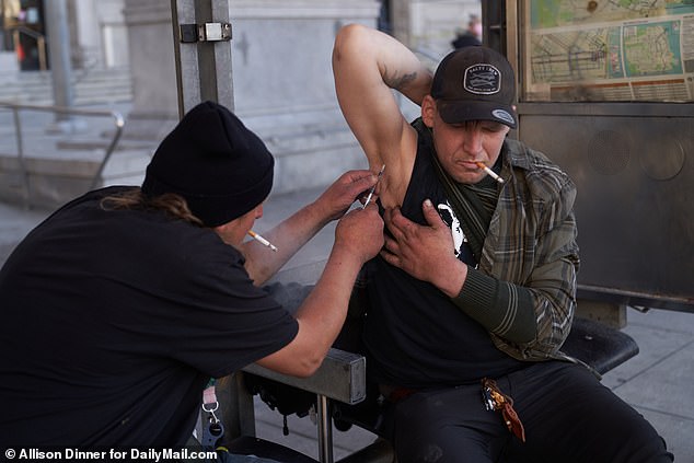 A homeless man injects fentanyl into his friend's armpit, due to a lack of usable veins, as people pass by San Francisco City Hall in September 2022.