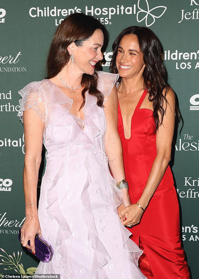 The two friends smile and hold hands in front of the cameras, with Meghan standing behind Kelly.