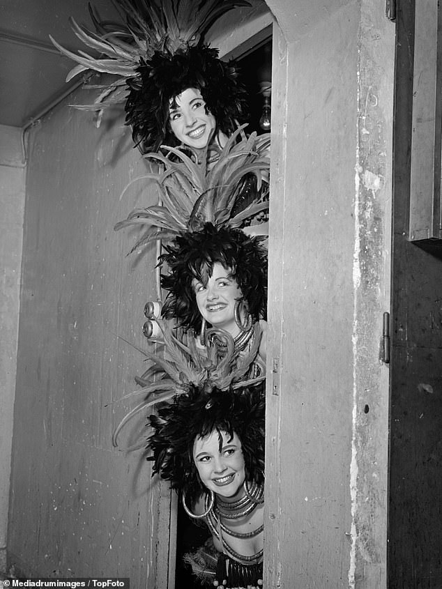 Three Bluebell Girls smile from their dressing room backstage at Le Lido in Paris in 1951. All three are wearing feather headdresses.