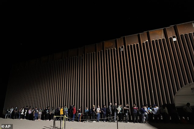 People line up against a border wall as they wait to apply for asylum after crossing the border from Mexico on Tuesday, July 11, 2023, near Yuma, Arizona. Thousands of migrants from the North African country of Mauritania have arrived in the United States in recent months, following a new route that takes them to Nicaragua and across the southern border.