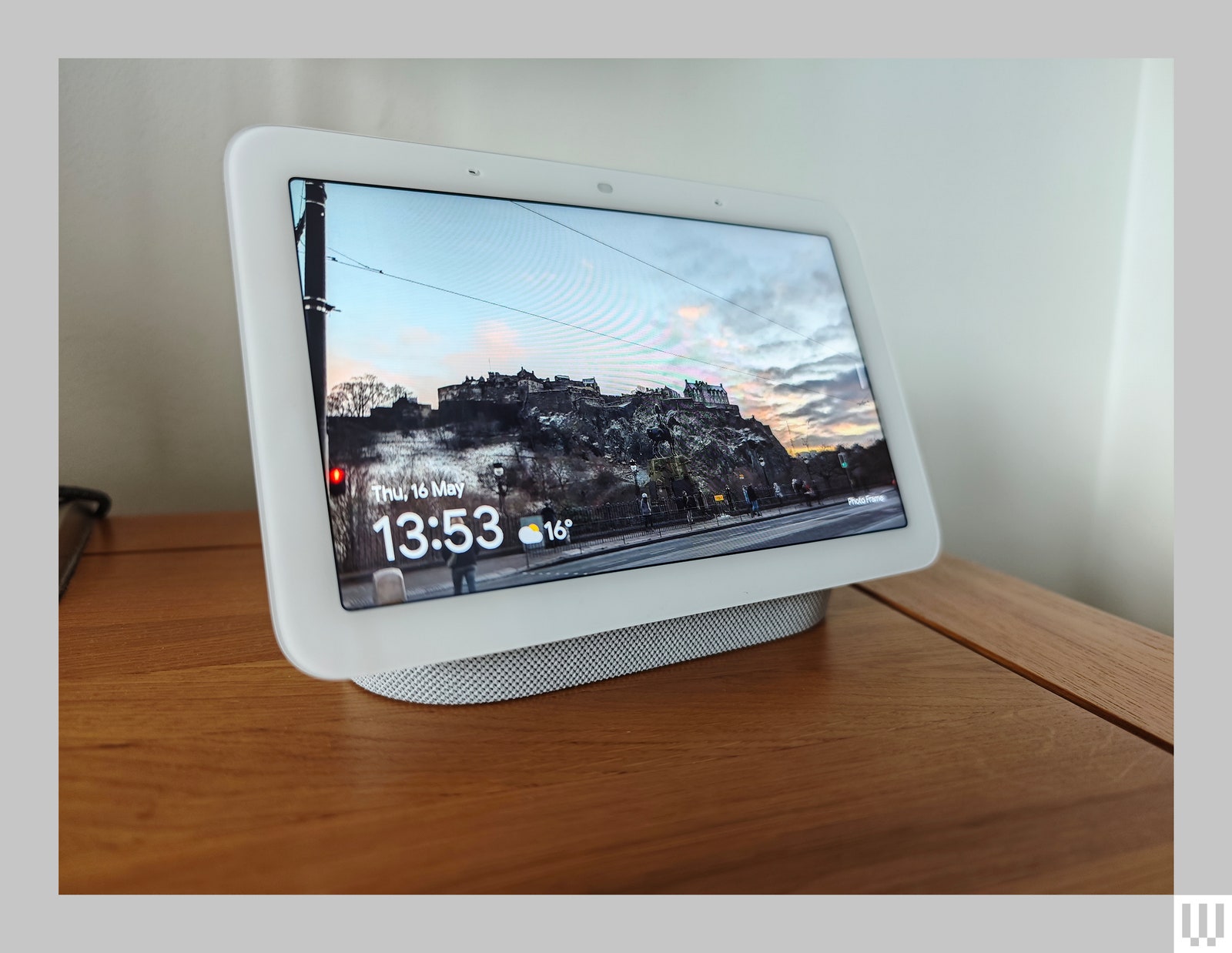 White tablet-like device placed on a wooden surface with a photo of buildings next to a cliff on the screen