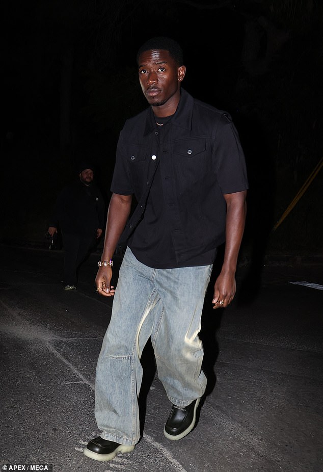 Damson Idris dressed smartly in an open short-sleeved black shirt over a plain T-shirt and a pair of gray jeans.