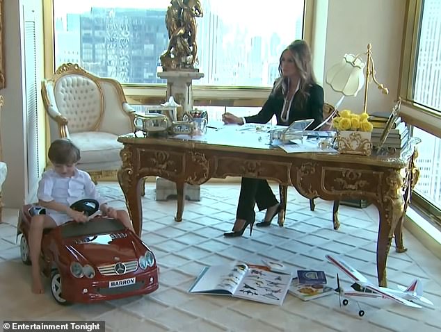 Melania sits at an ornate gold desk in a sleek black suit while her little one plays in his toy car.