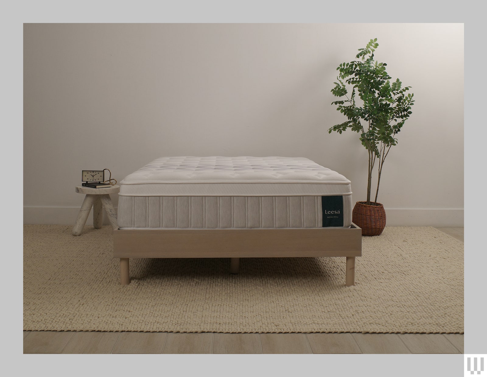 Leesa Sapira Chill a white mattress with tan trim on a minimalist wooden frame with a nightstand and a potted plant on...