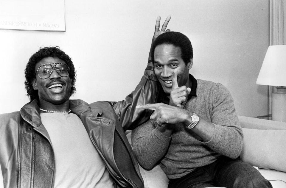 A black and white portrait of Eric Dickerson, left, and OJ Simpson smiling on a hotel couch.