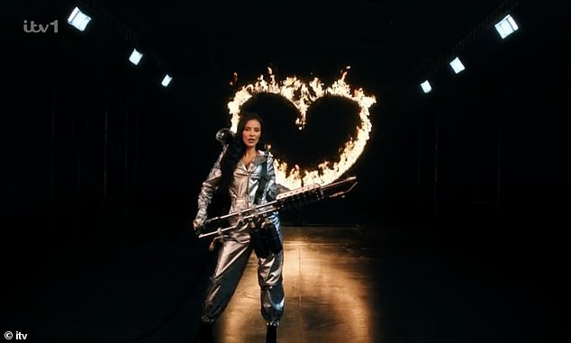 During the new teaser video, Maya appeared in a silver jumpsuit complete with a hood and a silver mask, wielding a flamethrower and lighting up a heart-shaped ring.