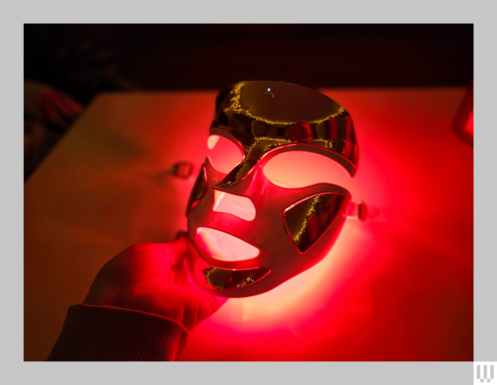 A dark room showing a hand holding a white and gold face mask backlit in red.