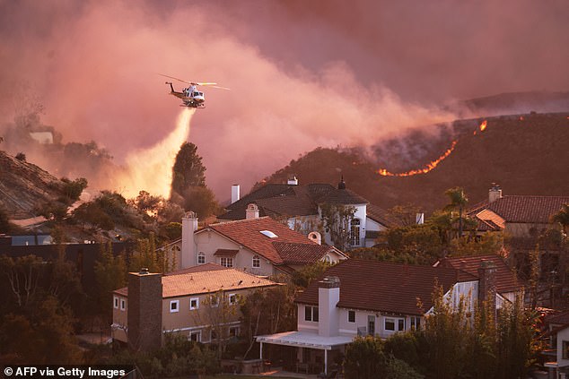 A helicopter drops water around homes threatened by the wind-driven Palisades Fire